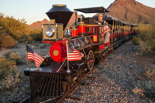 A vintage steam train with American flags, traveling through a desert landscape with mountains in the background.