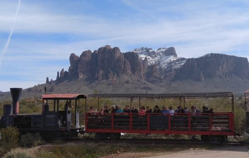 A vintage train with passengers travels through a desert landscape, with snow-capped mountains in the background.