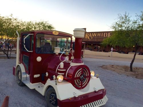 A vintage-style red and white train vehicle parked near a building labeled "Rawhide" under a clear sky.