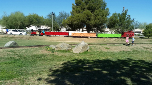 A small train with colorful cars travels through a grassy area, with children walking nearby and trees in the background.