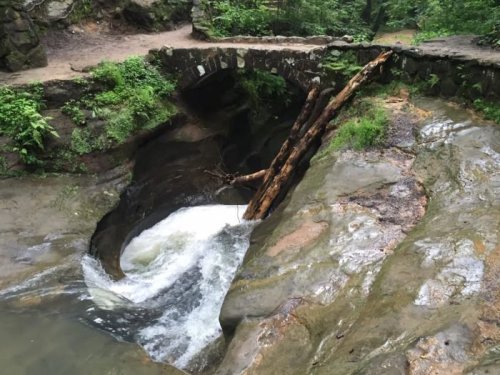 A small stone bridge over a rocky stream, with water flowing and logs resting on the rocks in a lush green forest.