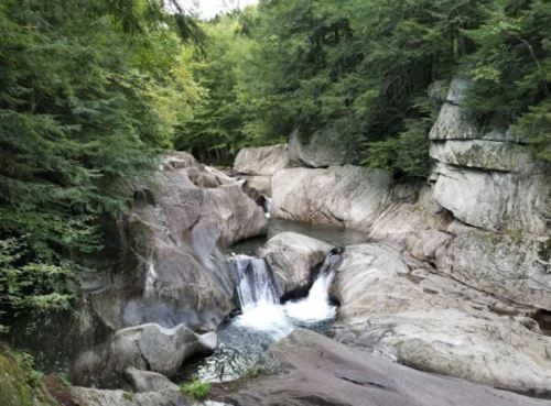 A serene landscape featuring a small waterfall cascading over rocky terrain, surrounded by lush green trees.
