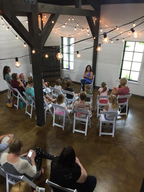 A group of children and adults seated in a cozy room, listening to a speaker under string lights.