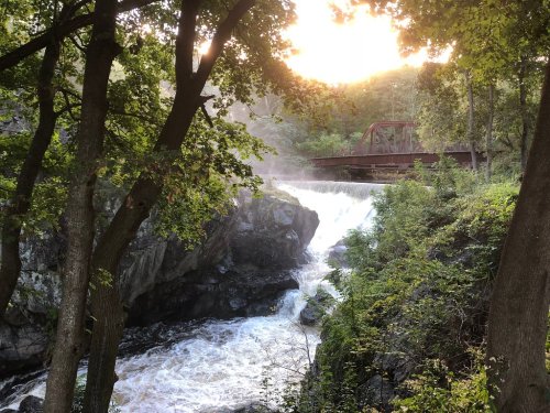 A waterfall cascades over rocks, framed by trees, with a bridge in the background and sunlight filtering through.
