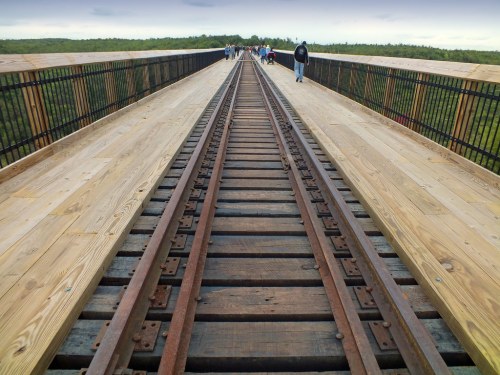 A wooden walkway alongside railway tracks stretches into the distance, with people walking on it against a cloudy sky.