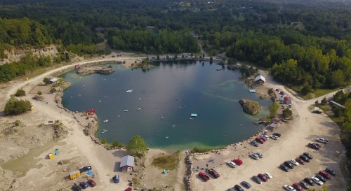 Aerial view of a circular lake surrounded by sandy shores, with people swimming and parked cars nearby.