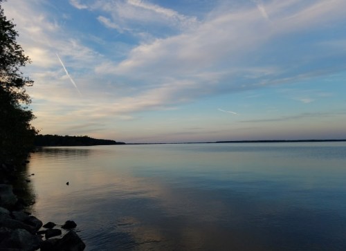 A serene lake at sunset, with calm waters reflecting the sky and soft clouds, framed by trees on the shore.