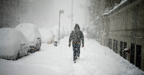 A person walks through a heavy snowstorm, with snow-covered cars and thick snowfall obscuring the surroundings.