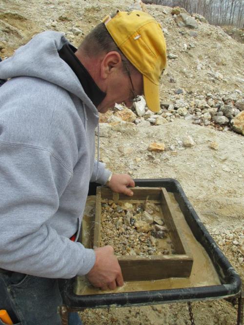 A man in a gray hoodie and yellow cap sifts through gravel in a wooden frame at a mining site.