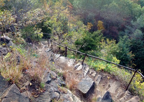 A winding stone path descends through lush greenery and autumn foliage, bordered by a metal railing.