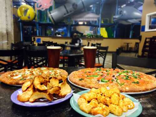 A table with two large pizzas, a plate of potato chips, and tater tots, set in a family-friendly restaurant.