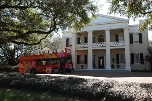 A red sightseeing bus parked in front of a large, historic white mansion surrounded by trees and greenery.