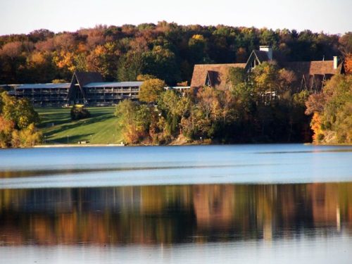 A serene lake reflects a wooded landscape and a large lodge with a sloped roof in the background. Autumn colors are visible.