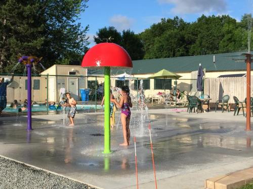 Children play in a splash pad with colorful water features, surrounded by a pool and lounge chairs on a sunny day.