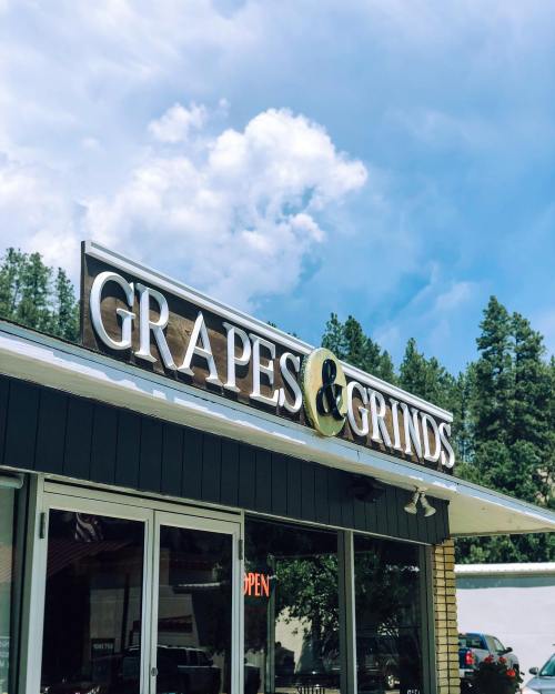 Sign for "Grapes & Grinds" storefront under a blue sky with clouds, surrounded by trees.
