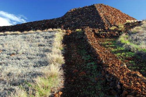 A rocky hill with a stone structure at the top, surrounded by grassy terrain under a blue sky.