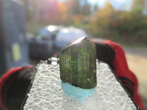 A green crystal held against a blurred background, reflecting sunlight with a sparkling effect.