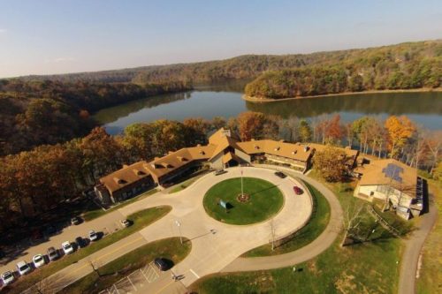 Aerial view of a lodge by a lake, surrounded by autumn trees and a circular driveway.