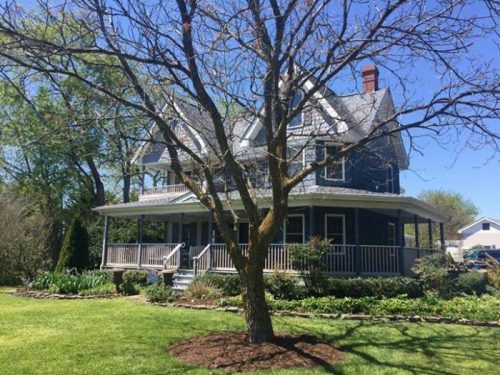 A large blue house with a porch, surrounded by green grass and trees on a sunny day.
