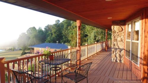 A wooden balcony with a table and chairs overlooks a scenic landscape at sunrise.