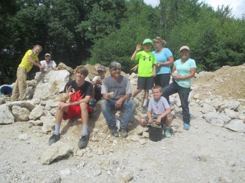 A group of children and adults pose together on a rocky site, smiling and holding tools, surrounded by greenery.