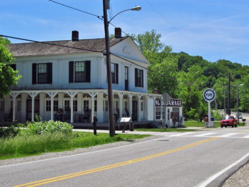 A vintage building with a porch, a garage sign, and a gas station sign, set along a rural road surrounded by greenery.