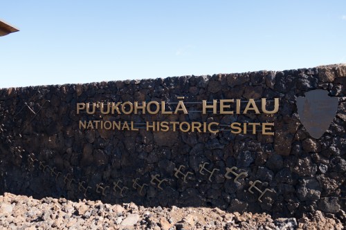 Sign for Pu'ukohola Heiau National Historic Site, featuring stone wall and bronze lettering against a clear blue sky.