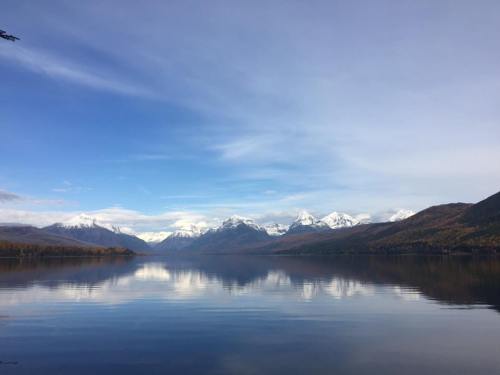 A serene lake reflects snow-capped mountains and a blue sky, surrounded by autumn-colored hills.