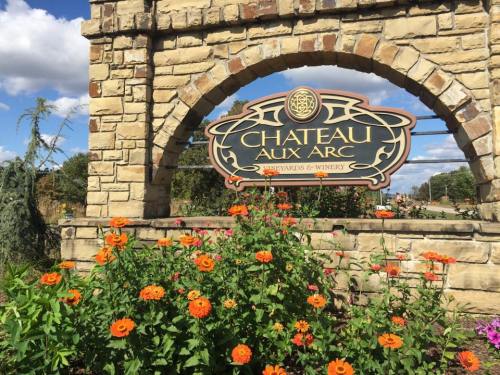 Sign for Chateau Aux Arc winery framed by colorful flowers and a stone archway under a blue sky.