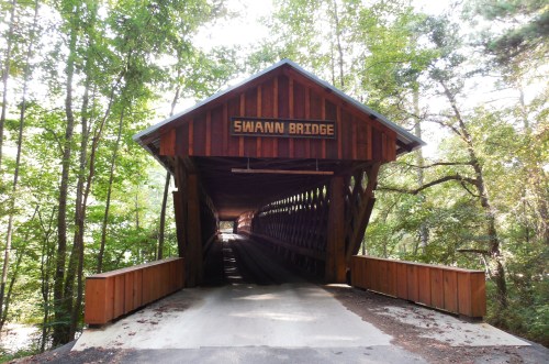 A wooden covered bridge named Swann Bridge, surrounded by lush green trees and a clear path leading inside.