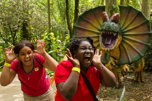 Two excited girls pose playfully with a colorful dinosaur statue in a lush, green environment.