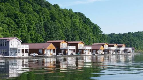 Row of lakeside cabins reflecting on calm water, surrounded by lush green hills under a clear blue sky.