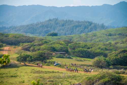 A group of people on horseback rides along a dirt path through lush green hills and mountains in the background.