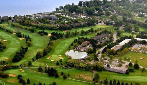 Aerial view of a lush green golf course with trees, a pond, and nearby residential areas along a lake.