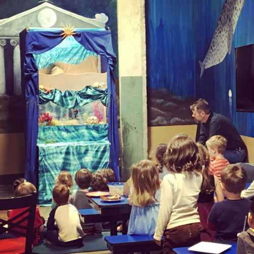 Children sit on benches watching a puppet show behind a colorful curtain in a playful, decorated room.