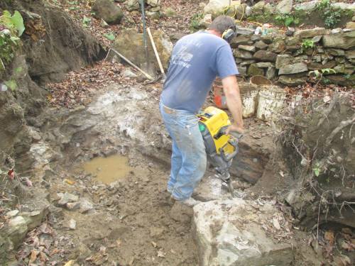 A person operates a jackhammer in a muddy excavation site surrounded by rocks and foliage.