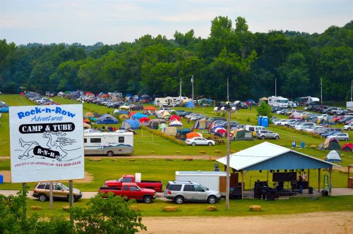 Aerial view of a campground with numerous tents and RVs, featuring a sign for Rock-n-Row Adventures Camp & Tube.