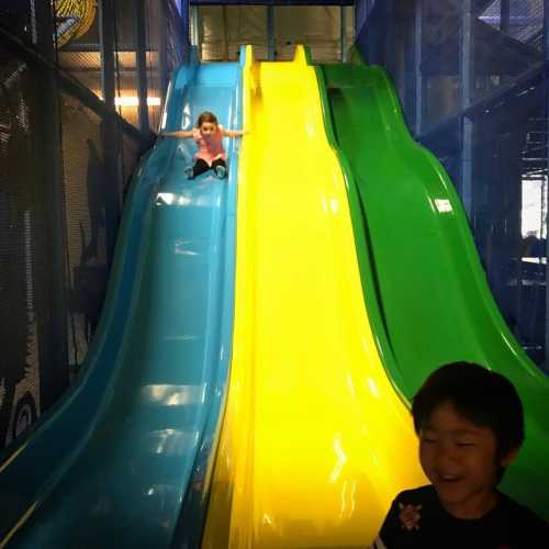 A child slides down a blue slide while another child smiles nearby on a colorful playground structure.