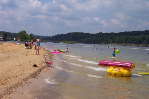 A sandy beach with people enjoying the water, floating on colorful inflatables under a partly cloudy sky.