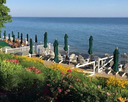 A serene lakeside view with green umbrellas, lounge chairs, and colorful flowers along a wooden deck by the water.