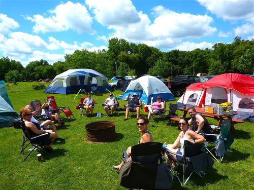 A group of people sitting around a campfire, surrounded by tents and green grass, enjoying a sunny day outdoors.
