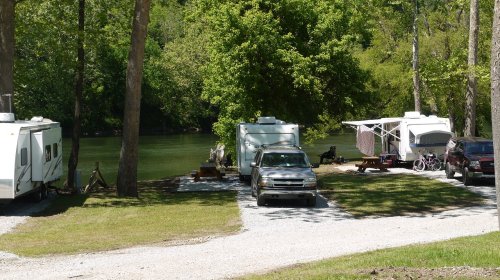 A serene campsite by a river, featuring parked RVs and trees in a lush green setting.