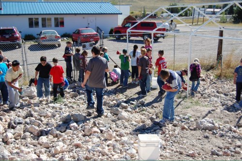 A group of children and adults are digging and exploring a rocky area outside a building on a sunny day.