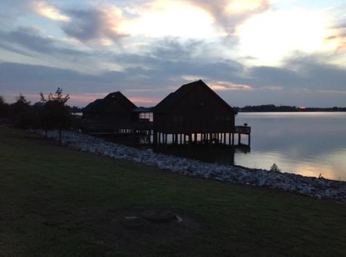 Two wooden cabins on stilts by a calm lake at sunset, with a cloudy sky and grassy shoreline.