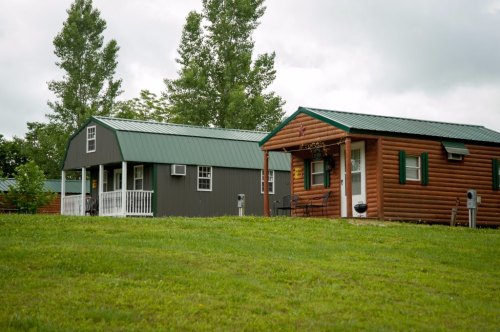 Two rustic cabins with green roofs sit on a grassy hillside, surrounded by trees under a cloudy sky.
