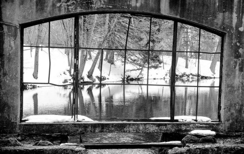 A broken window frame overlooks a snowy landscape and a calm, reflective river. Black and white photography.