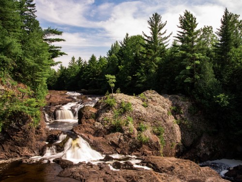 A serene waterfall cascades over rocky terrain, surrounded by lush green trees under a blue sky.
