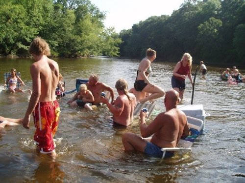 A group of people enjoying a summer day in a river, some sitting on chairs, others playing and relaxing in the water.
