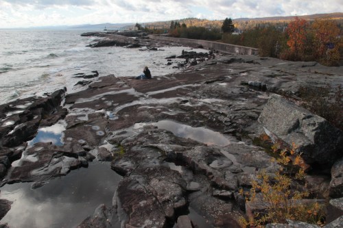 A rocky shoreline with a person sitting near the water, surrounded by autumn foliage and a cloudy sky.