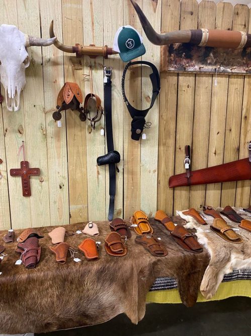 A display of leather goods, including belts and holsters, on a wooden wall with decorative items and a cowboy hat.